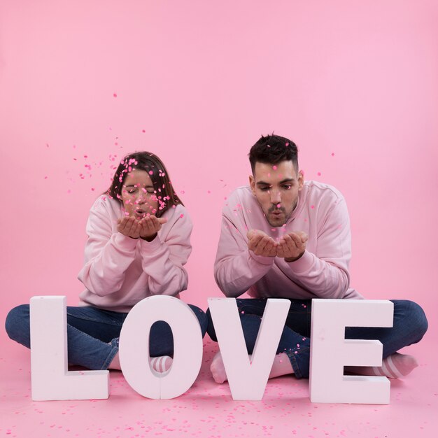 Jeune couple assis près de grand signe d&#39;amour et soufflant sur des confettis