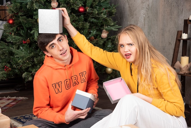 Photo gratuite jeune couple assis sur le sol avec des cadeaux et regardant la caméra près de l'arbre de noël