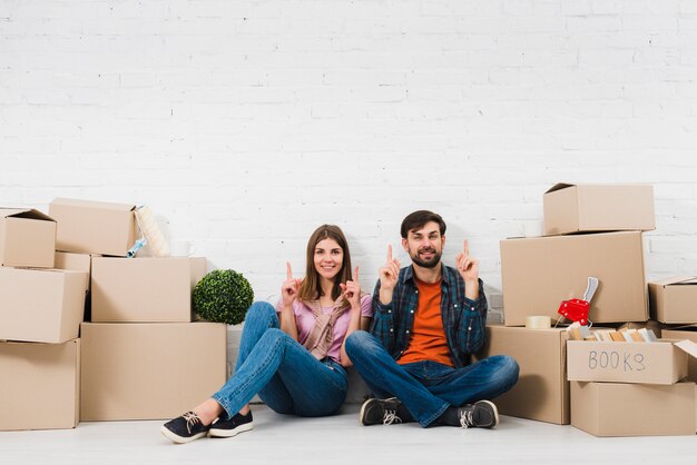 Jeune couple assis sur le sol, tenant leurs doigts vers le haut, assis entre les boîtes en carton