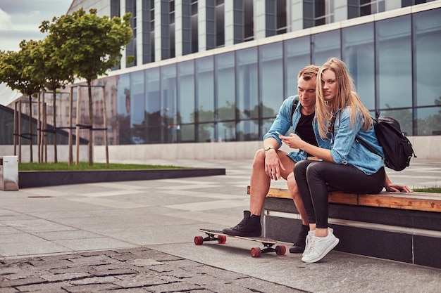 Jeune couple attrayant et heureux dans des vêtements décontractés assis sur un banc contre un gratte-ciel, regardant quelque chose sur le smartphone.