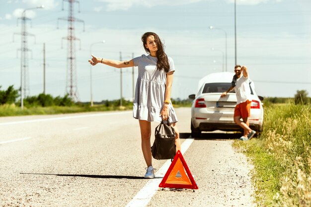 Le jeune couple a cassé la voiture alors qu'il se rendait au repos. Ils essaient d'arrêter les autres conducteurs et de demander de l'aide ou de faire de l'auto-stop. Relation, problèmes sur la route, vacances.