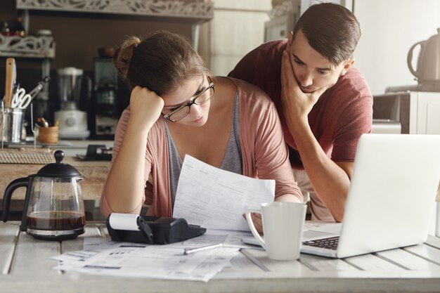 Jeune couple caucasien stressé face à des problèmes financiers, assis à la table de la cuisine avec des papiers, une calculatrice et un ordinateur portable et lisant un document de banque, l'air frustré et malheureux