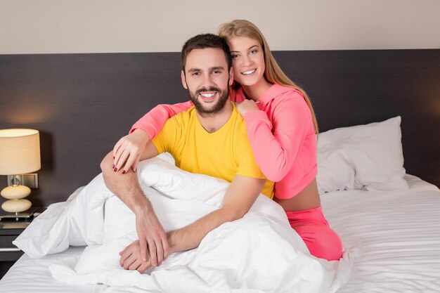 Jeune couple charmant allongé et s'amusant dans un lit, sourire heureux regardant la caméra, couvert sous couverture. Concept de famille, de coucher et de bonheur