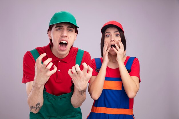 Jeune couple concerné en uniforme de travailleur de la construction et gars de casquette montrant les mains vides fille gardant les mains près du visage à la fois regardant la caméra isolée sur le mur blanc