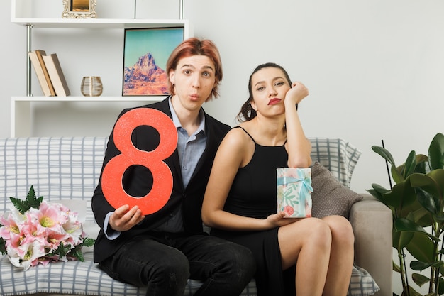 Photo gratuite jeune couple confus lors de la journée des femmes heureuses tenant le numéro huit avec une fille actuelle mettant la main sur la joue assise sur un canapé dans le salon