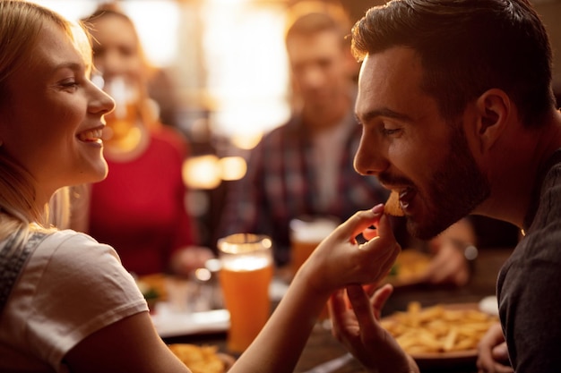 Photo gratuite jeune couple déjeunant avec leurs amis dans un restaurant jeune homme est nourri par sa petite amie avec des chips nacho