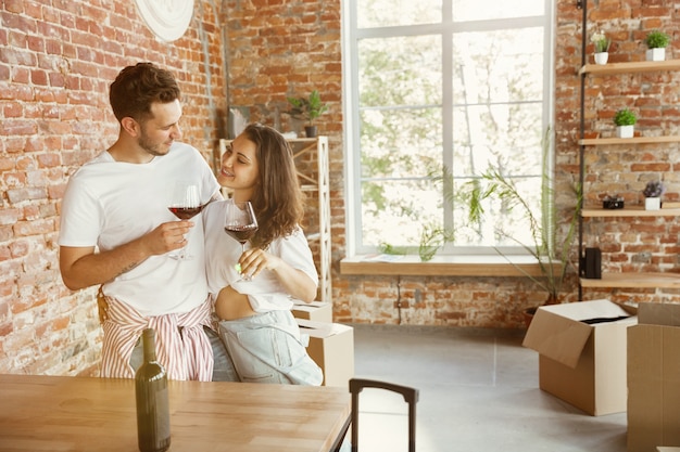 Photo gratuite un jeune couple a déménagé dans une nouvelle maison ou un nouvel appartement. boire du vin rouge, ranger et se détendre après le nettoyage et le déballage. ayez l'air heureux et confiant. famille, déménagement, relations, premier concept de maison.