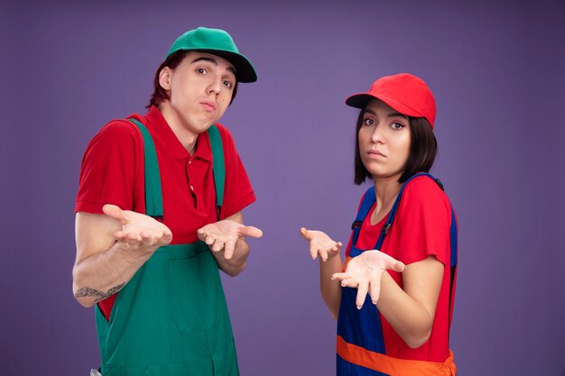 Jeune couple désemparé en uniforme de travailleur de la construction et casquette debout en vue de profil montrant les mains vides