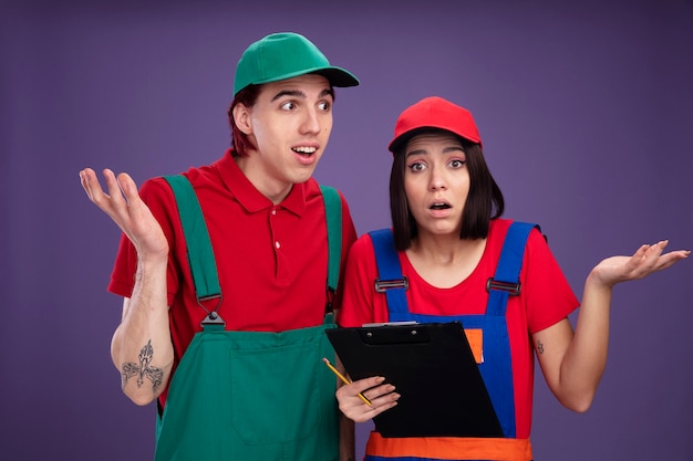 Jeune couple désemparé en uniforme de travailleur de la construction et fille de casquette tenant un crayon et un presse-papiers montrant tous les deux un gars à main vide regardant une fille de côté regardant la caméra isolée sur un mur violet