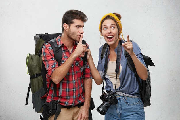 Jeune couple de deux randonneurs équipés d'accessoires touristiques et de sacs à dos profitant d'un voyage aventureux: homme barbu faisant signe chut avec le doigt, demandant à sa petite amie excitée de se taire