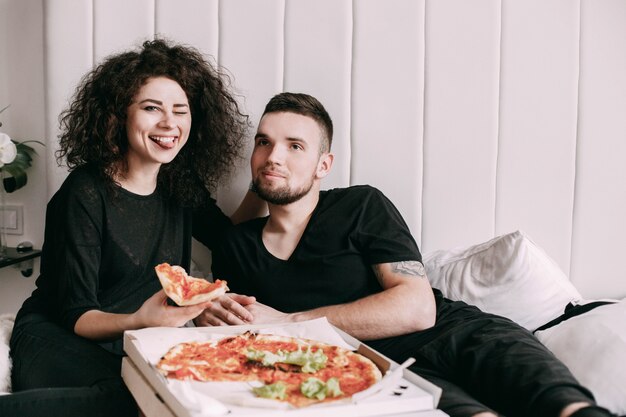 Un jeune couple drôle mange une pizza couchée sur le lit