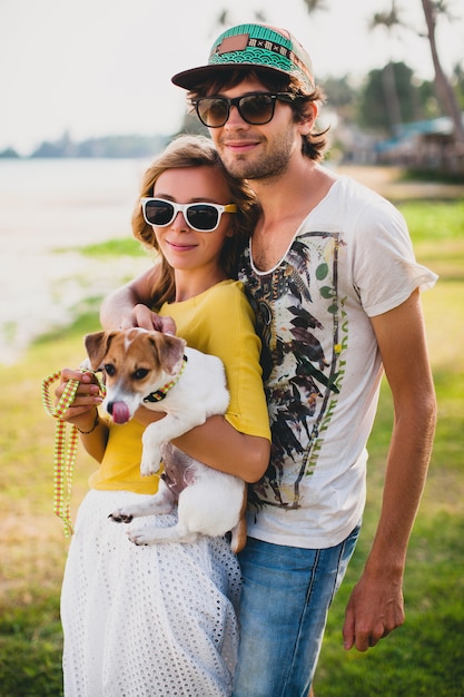 Jeune couple élégant hipster amoureux tenant un chien dans le parc tropical, souriant et s'amusant pendant leurs vacances, portant des lunettes de soleil, casquette, chemise jaune et imprimée