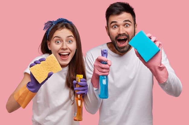 Un jeune couple de famille positif a des expressions faciales positives, utilise un spray de lavage chimique et une éponge pour nettoyer la fenêtre dans la chambre