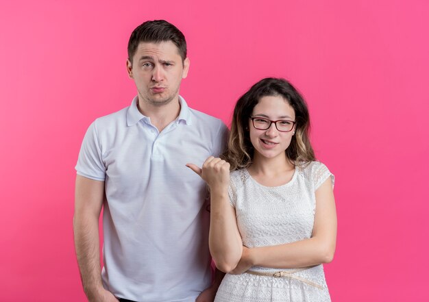 Jeune couple femme pointant avec le doigt sur son petit ami confus debout sur un mur rose