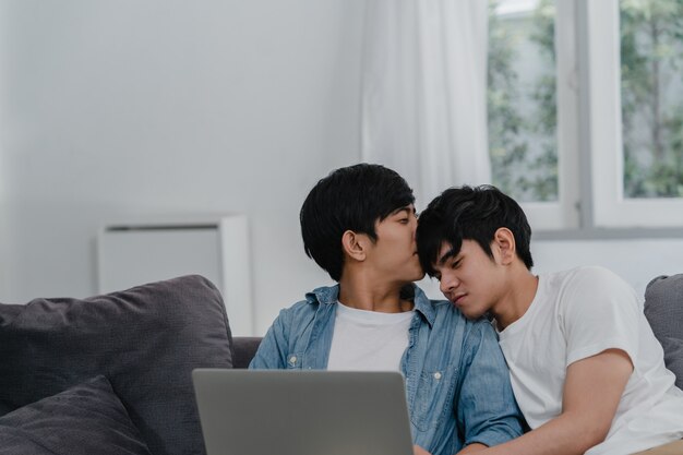 Jeune couple gay à l'aide d'un ordinateur portable à la maison moderne. Les LGBTQ + asiatiques heureux se détendent en utilisant la technologie en regardant un film sur Internet tout en se trouvant dans le canapé de la maison.