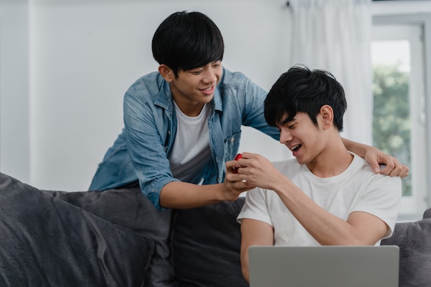 Jeune couple gay asiatique propose à la maison moderne, les hommes LGBTQ coréens adolescents souriants heureux ont un temps romantique tout en proposant et alliance surprise porter bague de mariage dans le salon à la maison.