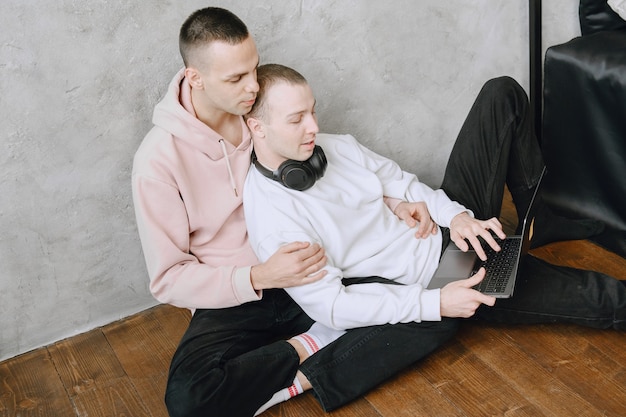 Jeune couple gay assis sur le sol à l'aide d'un ordinateur portable, à l'aide d'un casque, écouter de la musique ensemble, s'étreindre