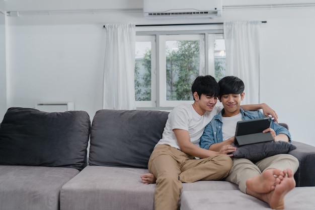 Jeune couple gay avec tablette à la maison. Les hommes LGBTQ + asiatiques heureux se détendent en utilisant la technologie en regardant un film sur Internet tout en se trouvant dans le canapé.