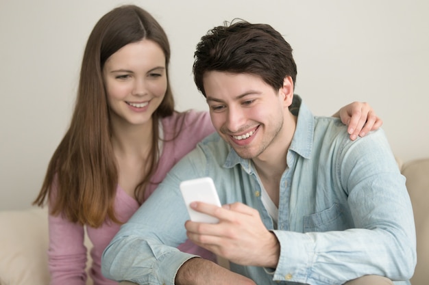 Jeune couple heureux assis sur un canapé à la maison à l&#39;aide de smartphone