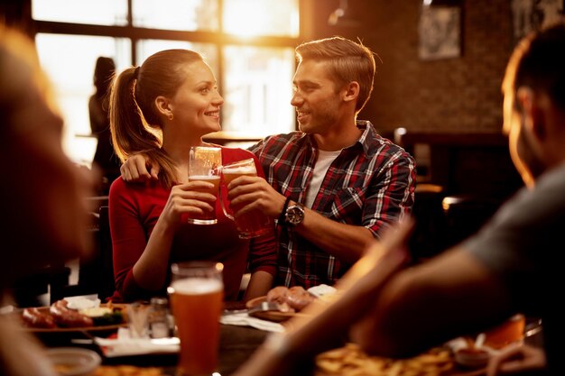 Jeune couple heureux portant un toast avec de la bière et célébrant tout en déjeunant avec leurs amis dans un pub