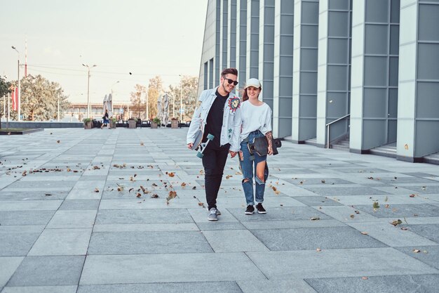 Un jeune couple heureux se tient la main et marche avec des planches à roulettes dans une rue moderne par temps venteux.