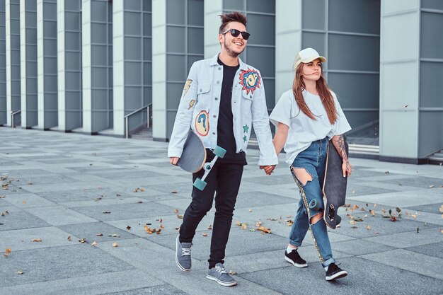 Un jeune couple heureux se tient la main et marche avec des planches à roulettes dans une rue moderne par temps venteux.