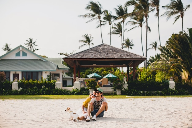 Jeune couple hipster élégant dans l'amour à pied jouant chiot chien Jack Russell, plage tropicale, tenue cool, ambiance romantique, s'amuser, ensoleillé, homme femme ensemble, horizontal, vacances, maison maison villa