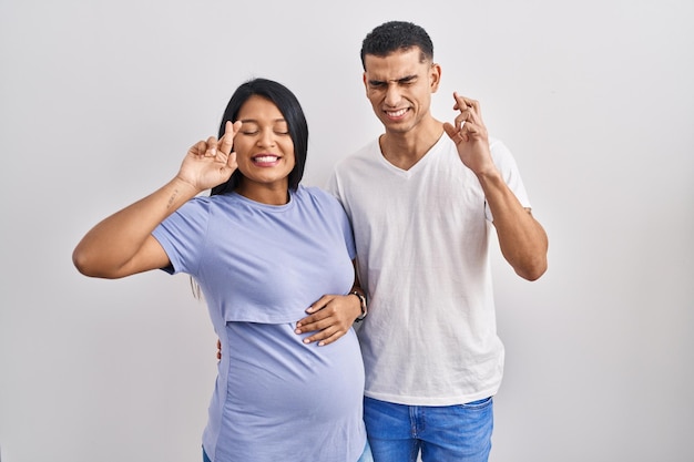 Photo gratuite jeune couple hispanique attend un bébé debout sur fond gesticulant doigt croisé souriant avec espoir et les yeux fermés chance et concept superstitieux