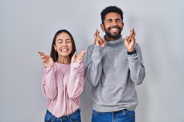 Jeune couple hispanique debout ensemble gesticulant doigt croisé souriant avec espoir et les yeux fermés chance et concept superstitieux