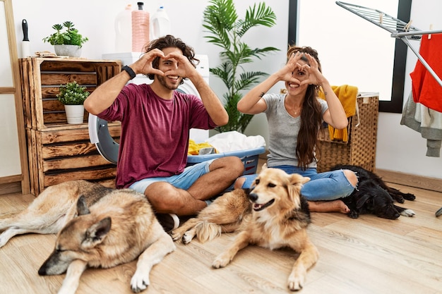 Jeune couple hispanique faisant la lessive avec des chiens faisant la forme de coeur avec la main et les doigts souriant regardant à travers le signe