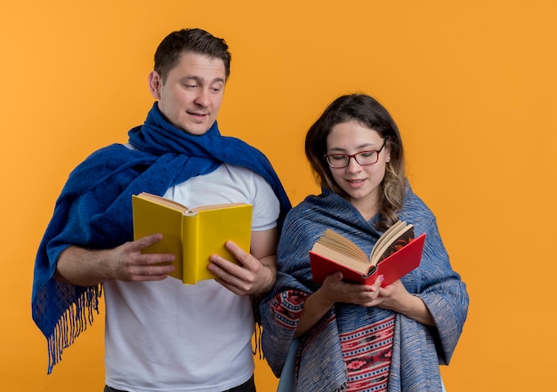 Jeune couple homme et femme avec des couvertures tenant des livres heureux et positif souriant debout sur un mur orange