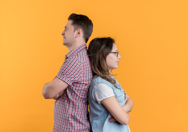 Jeune couple homme et femme dans des vêtements décontractés debout dos à dos après combat debout sur un mur orange