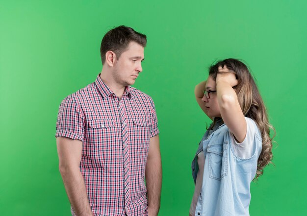 Jeune couple homme et femme dans des vêtements décontractés a déplu à l'homme avec une expression triste en regardant sa petite amie confuse debout sur le mur vert