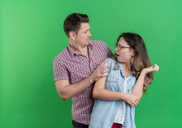 Jeune couple homme et femme dans des vêtements décontractés homme confus demandant à sa petite amie en colère pour le pardon debout sur le mur vert