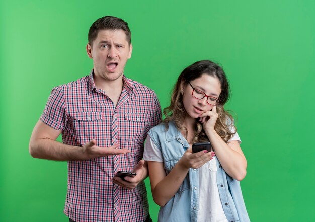 Jeune couple homme et femme dans des vêtements décontractés homme confus pointant avec le bras sur sa fille occupée qui parle au téléphone mobile debout sur le mur vert