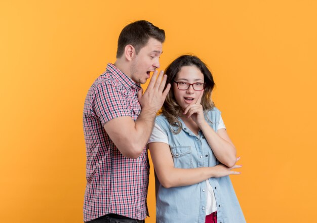 Jeune couple homme et femme dans des vêtements décontractés homme surpris en chuchotant un secret à sa petite amie sur orange