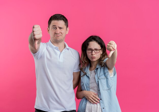 Jeune couple homme et femme dans des vêtements décontractés mécontents montrant les pouces vers le bas debout sur un mur rose