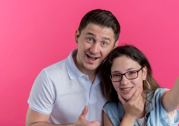 Jeune couple homme et femme dans des vêtements décontractés prenant selfie homme souriant heureux et positif montrant les pouces vers le haut sur rose