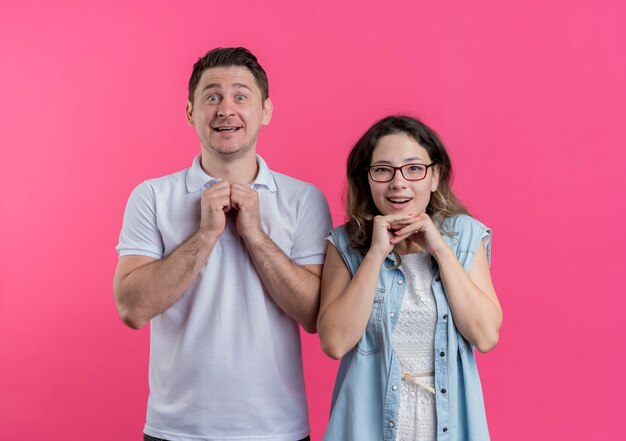 Jeune couple homme et femme dans des vêtements décontractés se tenant la main ensemble en attente émotionnelle de surprise debout sur un mur rose