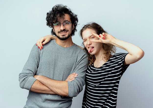 Jeune couple homme et femme femme heureuse et positive stiking out tongue montrant le signe de la victoire debout sur un mur blanc