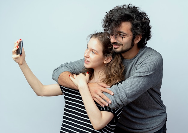 Jeune couple homme et femme heureux en amour, femme heureuse de prendre une photo d'eux à l'aide de smartphone debout sur un mur blanc