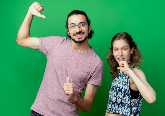 jeune couple homme et femme, heureux et positif pointant sur eux-mêmes debout sur un mur végétal