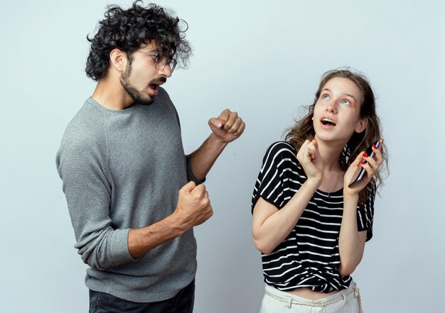 Jeune couple homme et femme, homme déçu avec les poings serrés regardant fille surprise qui tient debout smartphone sur fond blanc