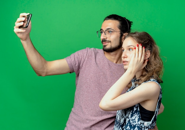 Jeune couple homme et femme, homme heureux de prendre une photo d'eux à l'aide de son smartphone debout sur un mur vert