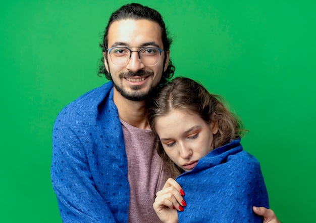 Jeune couple homme et femme, homme heureux serrant sa petite amie bien-aimée en souriant tout en l'enveloppant dans une couverture chaude debout sur fond vert