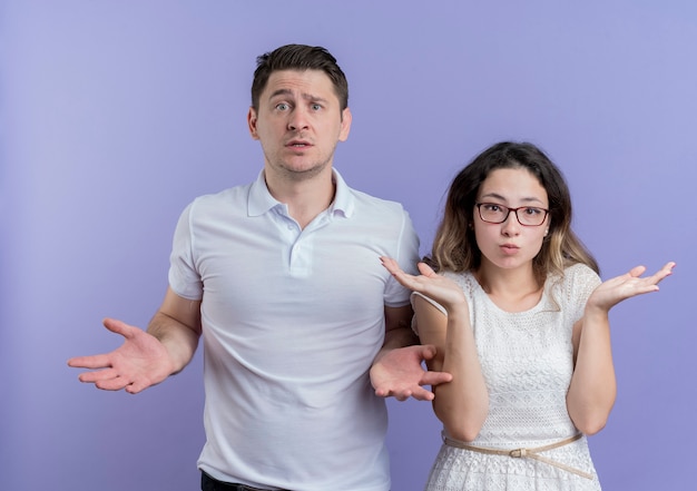 Jeune couple homme et femme regardant la caméra confus étalant les bras sur les côtés n'ayant pas de réponse debout sur le mur bleu