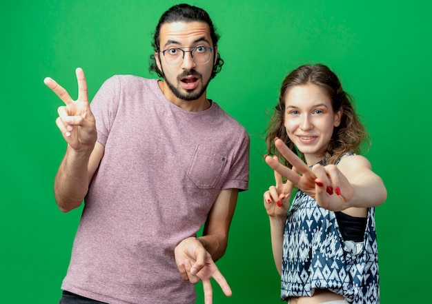 Jeune couple homme et femme regardant la caméra en souriant montrant le signe de la victoire debout sur fond vert