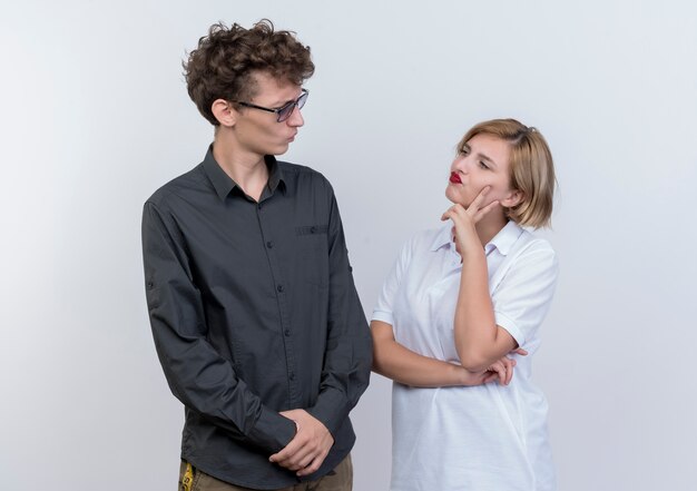 Jeune couple homme et femme se regardant avec une expression sceptique debout sur un mur blanc