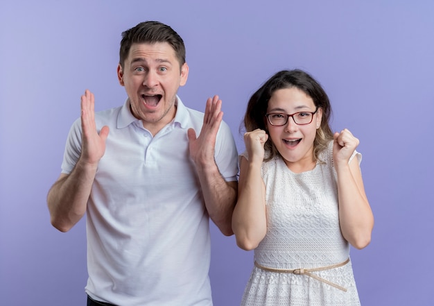 Jeune couple homme et femme serrant les poings heureux et excité debout sur le mur bleu