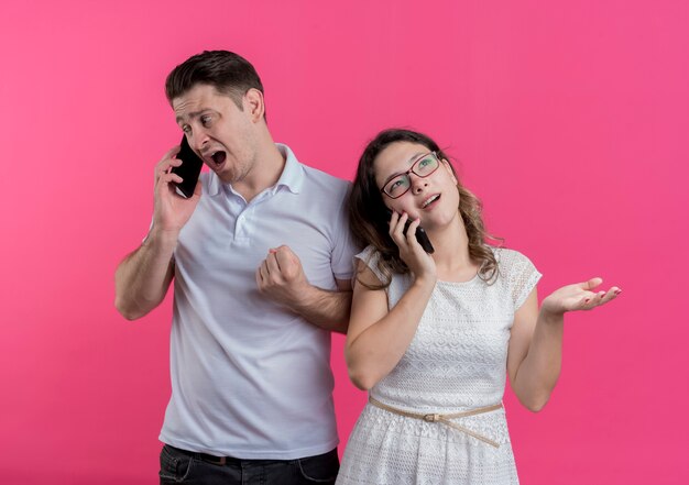 Jeune couple homme et femme en vêtements décontractés parlant sur les téléphones mobiles mécontent et confus debout sur le mur rose